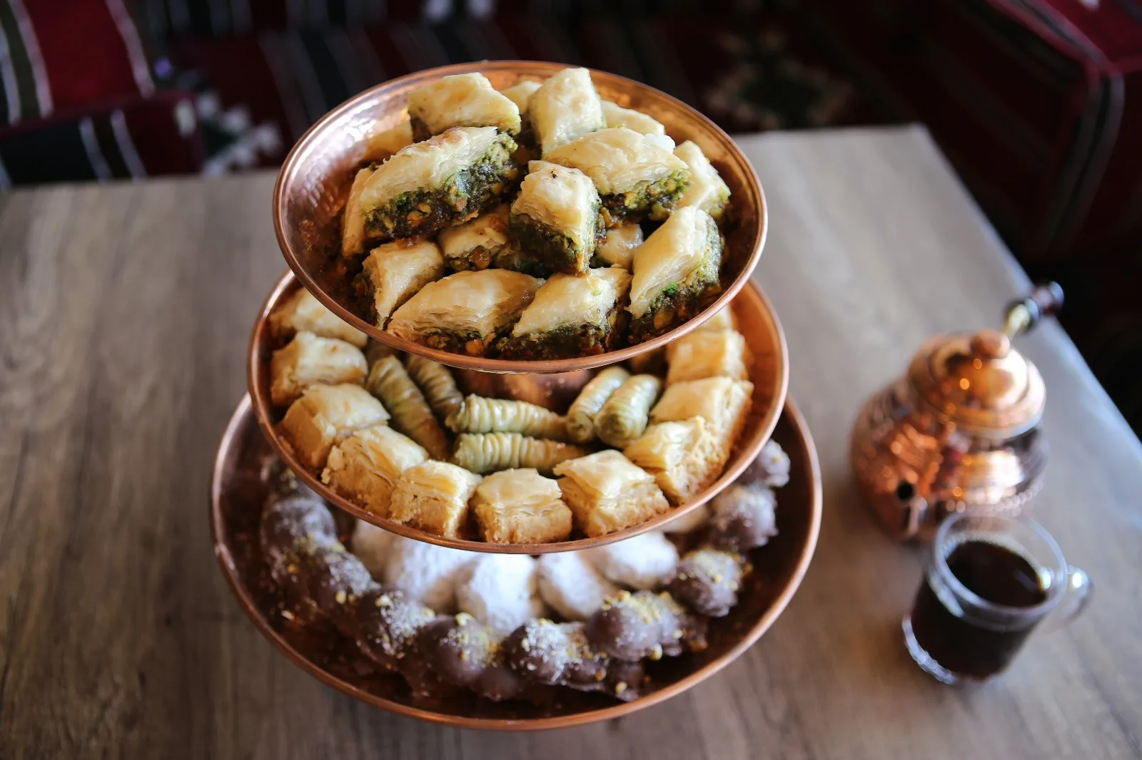 A three-tier copper stand of mixed Iraqi sweets served with a small pot of qahwa